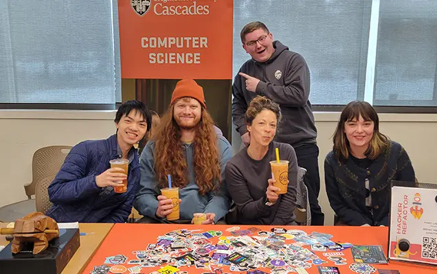 Students posing with a table full of stickers. 3 of them are holding drinks. 
