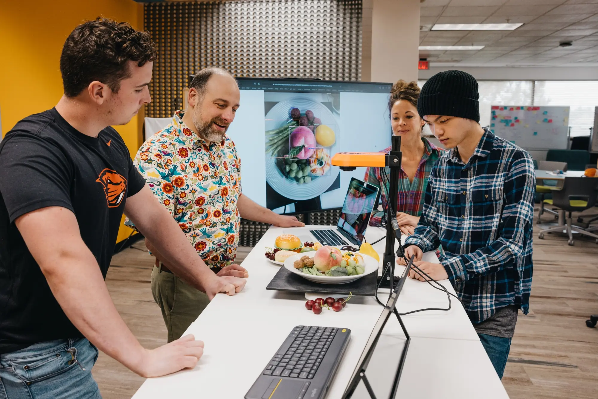 Group of people standing at table with computer, lab equipment and a plate of food.